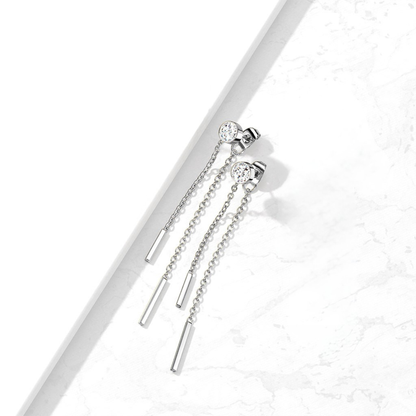 A pair of silver-toned dangle earrings with clear round gemstones and elongated cylindrical and bead chain drops, photographed on a white marble surface with a white geometric prop.